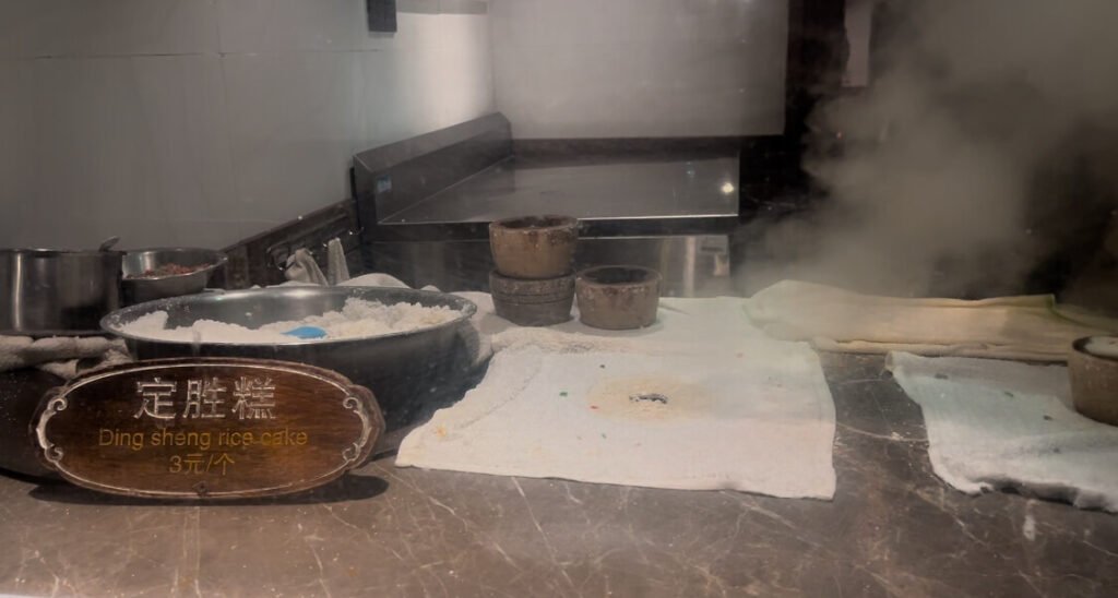 중국 전통 음식 중 하나인 'Ding sheng rice cake'라고 적혀있는 문구와 함께 솥 안에 쌀이 담겨 있으며 그 주위에 하얀 천 들이 테이블 위에 있습니다. 그 뒤에는 쌀을 담는 틀이 보이며 오른쪽에는 연기가 나고 있습니다. 우전마을 먹거리 중 하나입니다.